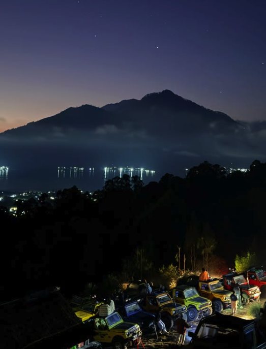 Jeeps parked before dawn at the Mount Batur viewpoint, with village lights and fishing boats on Lake Batur below