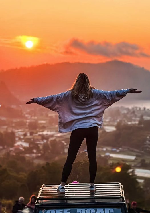 A traveller standing on a jeep roof rack with arms out, looking across the Batur caldera at first light