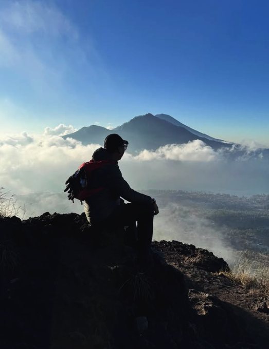 A hiker resting on the Mount Batur crater rim with Agung and Abang rising through the cloud