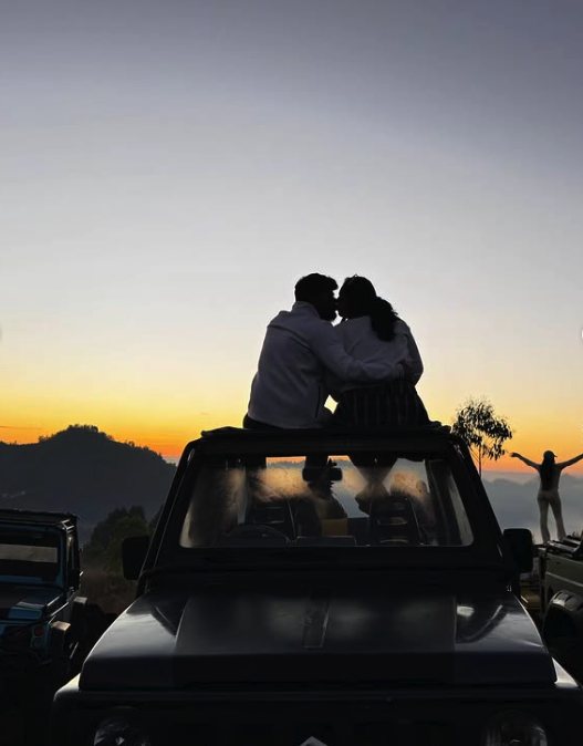 Two travellers silhouetted on a jeep roof at dawn, with other jeeps drawn up along the viewpoint