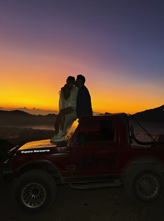 A couple on the bonnet of a red jeep against an orange and purple dawn over the caldera