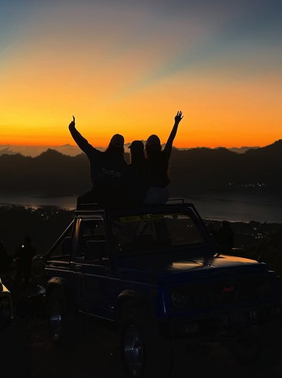 Three friends sitting on a jeep roof with their arms raised as the sun clears the ridge above Lake Batur