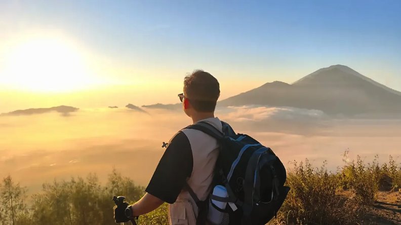 A walker on the Mount Batur summit ridge at sunrise, above a sea of cloud with Mount Agung on the horizon