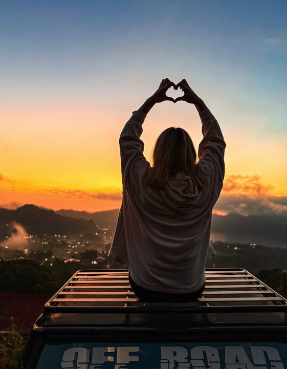 A traveller on a jeep roof making a heart shape against the sunset over the Batur caldera