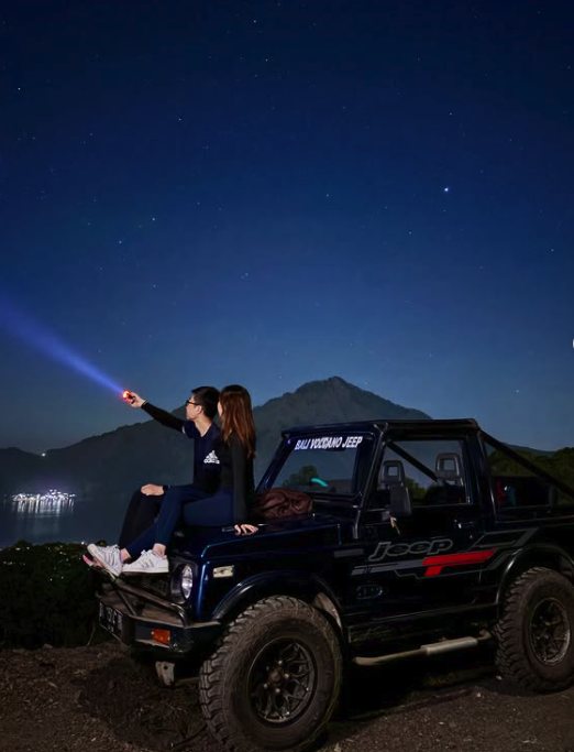 Two travellers on a jeep bonnet under the stars before dawn, a torch beam pointing at the Mount Batur cone